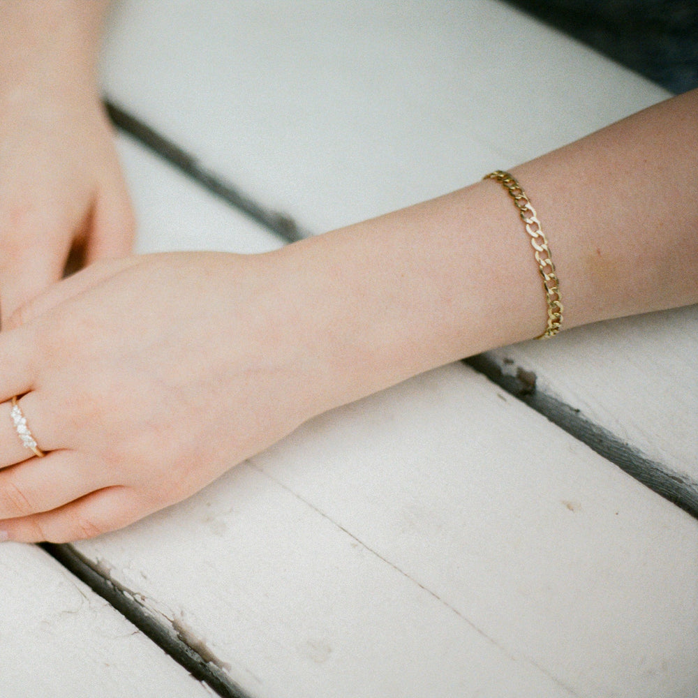 
                      
                        Close-up of a person's hands resting on a white wooden surface. One hand has a plain ring on the fourth finger, while the other wrist is adorned with the Bold Curb Bracelet by Pear., a thin gold chain perfect for everyday wear. The background is slightly blurred, focusing attention on the hands and jewelry.
                      
                    