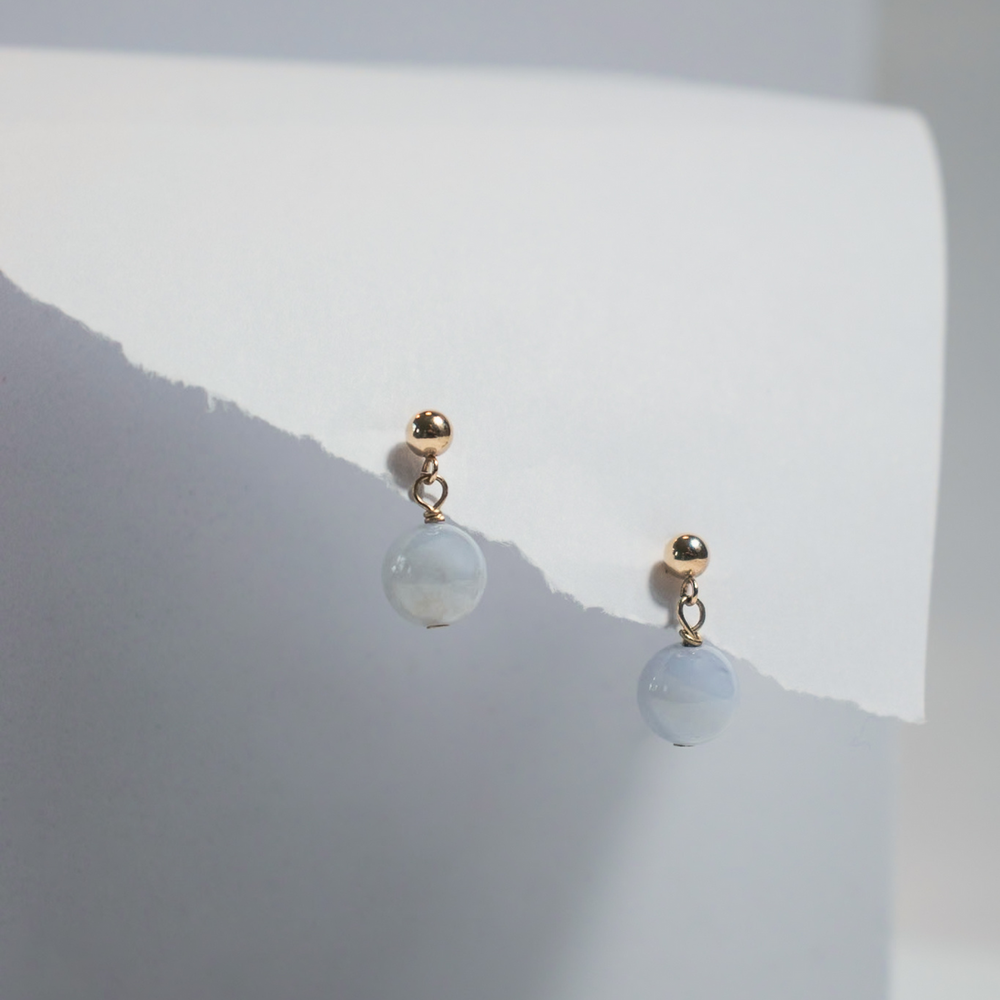 
                      
                        The Agate Boule Earrings by Pear. are a pair of golden stud earrings featuring small natural agate ball pendants. They are displayed against a white, folded paper background, showcasing a minimalistic and elegant style.
                      
                    