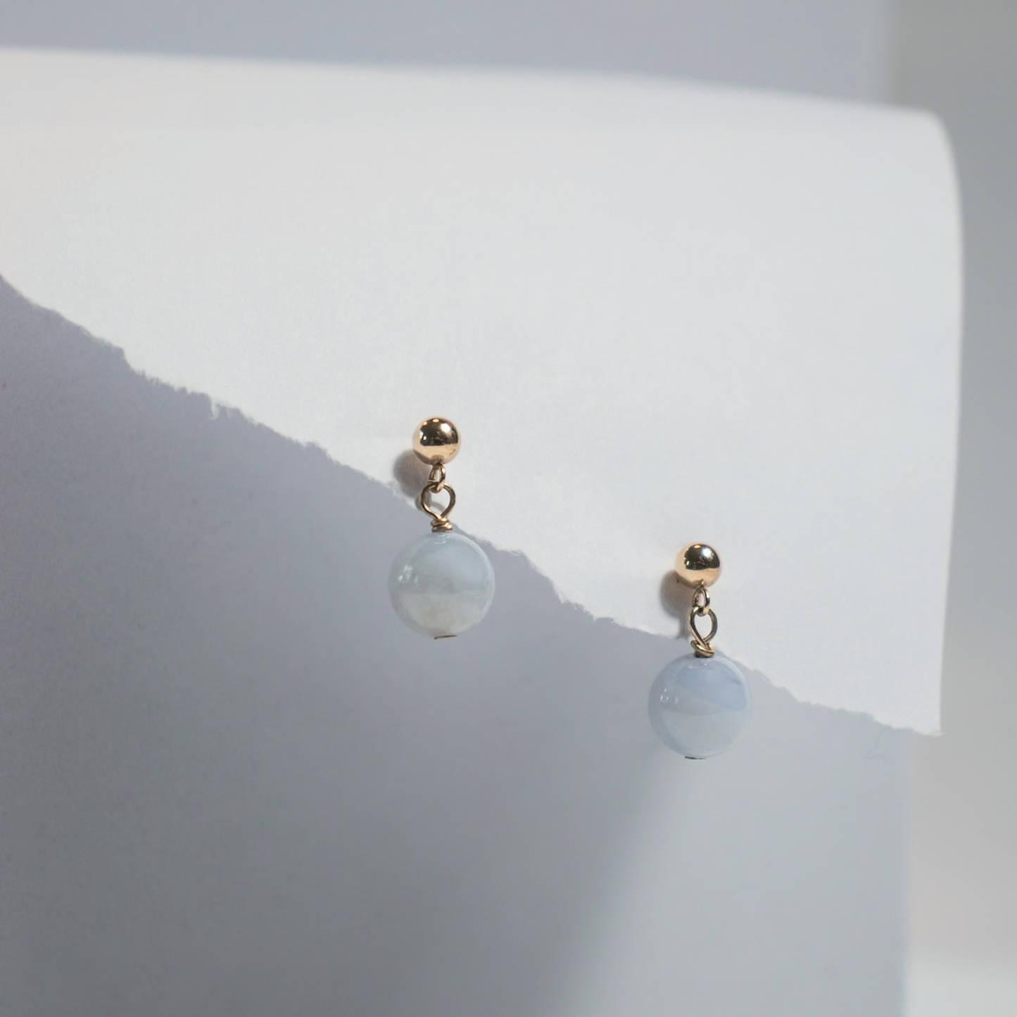 The Agate Boule Earrings by Pear. are a pair of golden stud earrings featuring small natural agate ball pendants. They are displayed against a white, folded paper background, showcasing a minimalistic and elegant style.