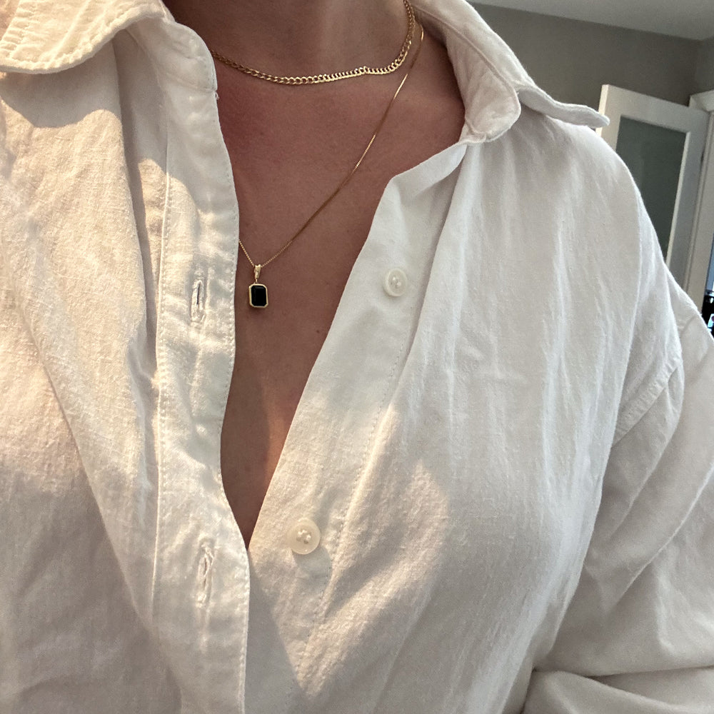 A person wearing a white button-up shirt, with the top few buttons undone, revealing two gold necklaces with pendants. The background shows a lightly lit indoor room with minimal decor.