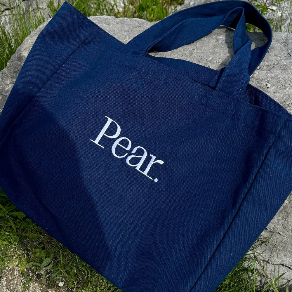 
                      
                        A dark blue Pear. Everyday Tote Bag with the brand name "Pear." printed in white lies on a rocky ground, surrounded by green grass and plants. The cotton canvas tote bag features two handles and appears to be made of sturdy, heavier weight fabric.
                      
                    