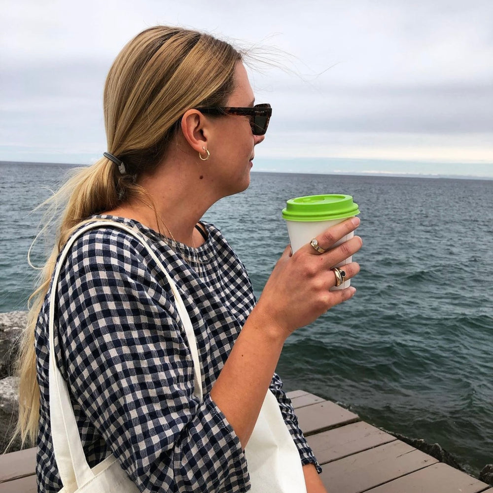 A person with long blond hair, wearing sunglasses and a black-and-white checkered shirt, stands on a wooden pier holding a white cup with a green lid. The person has a white tote bag over their shoulder and is gazing out at the ocean. The sky is cloudy.