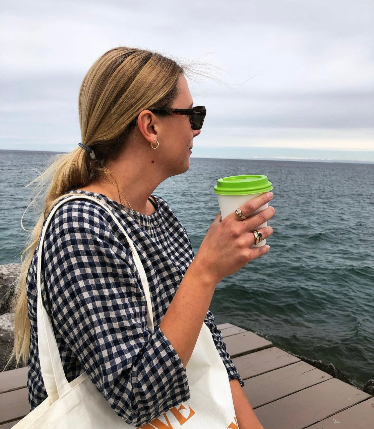 A person with long blond hair, wearing sunglasses and a black-and-white checkered shirt, stands on a wooden pier holding a white cup with a green lid. The person has a white tote bag over their shoulder and is gazing out at the ocean. The sky is cloudy.