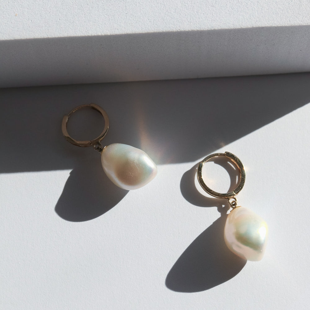 A pair of gold hoop earrings each adorned with a single dangling white pearl. The earrings are placed on a white surface with a shadow cast by light coming from the left side of the image. The pearls have a soft, iridescent glow.