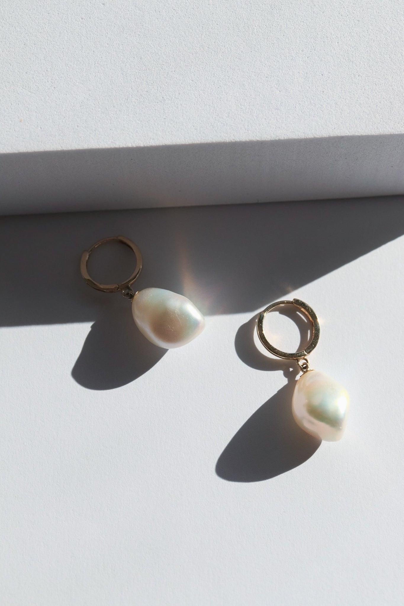 A pair of gold hoop earrings each adorned with a single dangling white pearl. The earrings are placed on a white surface with a shadow cast by light coming from the left side of the image. The pearls have a soft, iridescent glow.