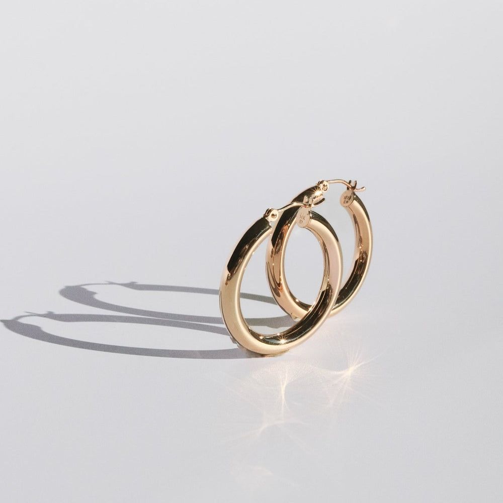 
                      
                        A pair of Pear. Thick Tube Hoops, crafted from solid 14K gold with smooth, polished surfaces, are placed on a light grey background. The lightweight hoops are slightly overlapping, casting soft shadows on the surface below them. The gold has a reflective quality that creates subtle highlights.
                      
                    