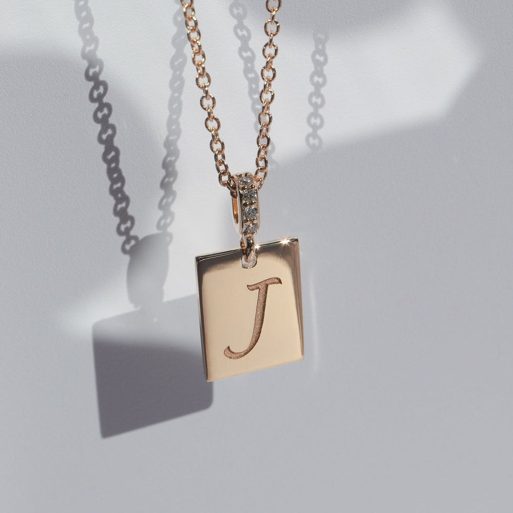 
                      
                        A Diamond Cleo Initial Necklace by Pear, featuring a square pendant with an engraved letter "J." The customizable gold pendant is adorned with small gemstones along the bail, and its shadow is cast on a light gray surface.
                      
                    