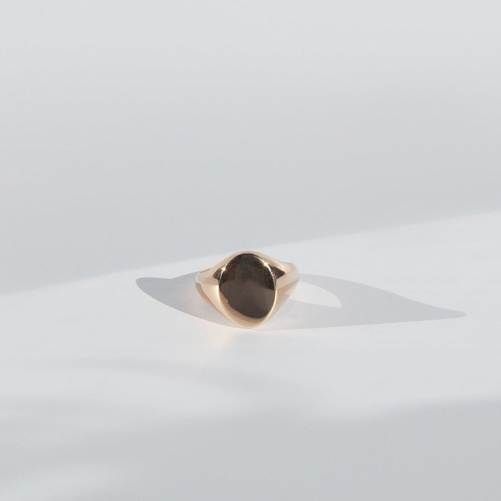 
                      
                        The Pear Oval Signet Ring, crafted from solid gold with an oval-shaped, smooth, and polished face, rests on a light surface, casting a subtle shadow. The soft lighting underscores the ring's simple and elegant design.
                      
                    