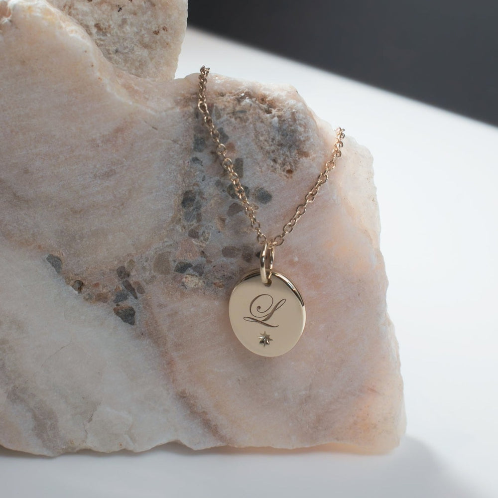 
                      
                        An oval engravable necklace from Pear rests on a marble surface, featuring a round, engraved "L" and a small star. The pendant hangs from an adjustable cable chain, creating a minimalist look with soft lighting.
                      
                    