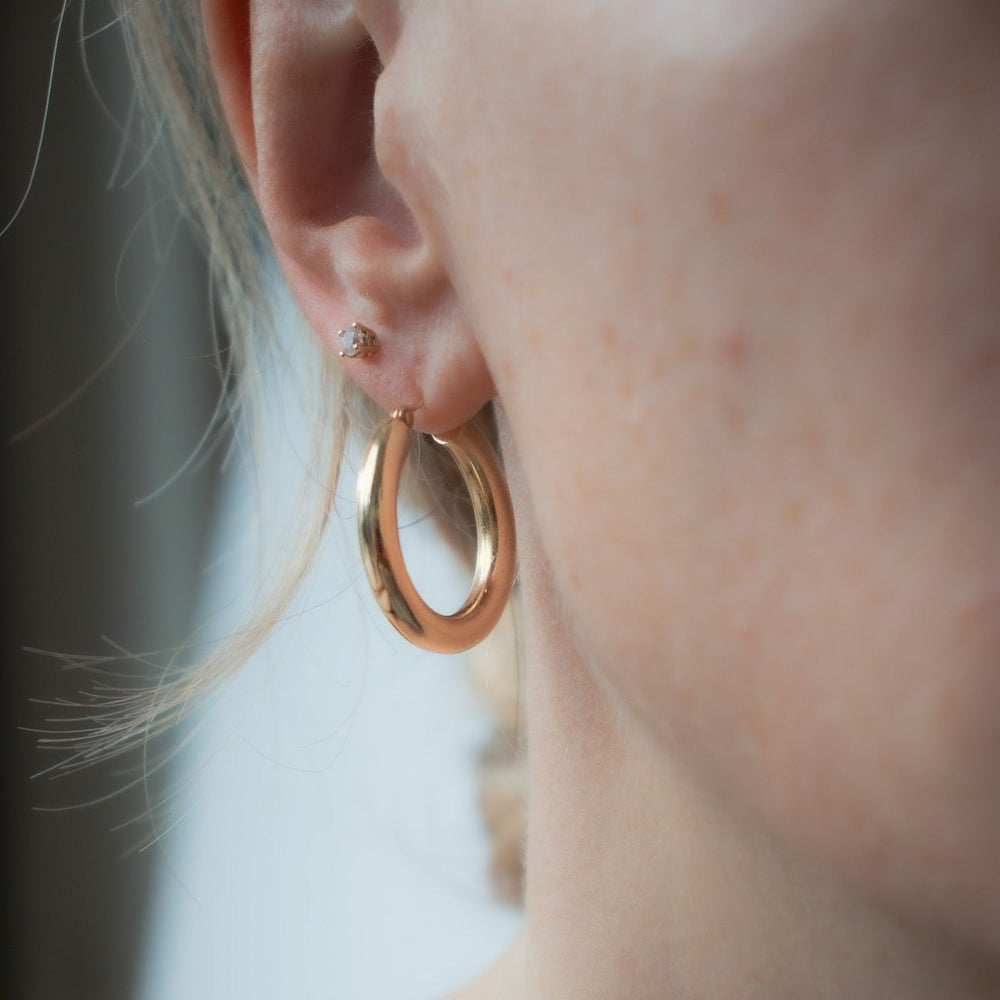 
                      
                        Close-up of a person's ear adorned with lightweight earrings, including a small stud and a large solid 14K gold hoop from Pear.'s Thick Tube Hoops collection. The image focuses on the jewelry, highlighting the earlobe and part of the person's neck and cheek. The background is softly blurred.
                      
                    