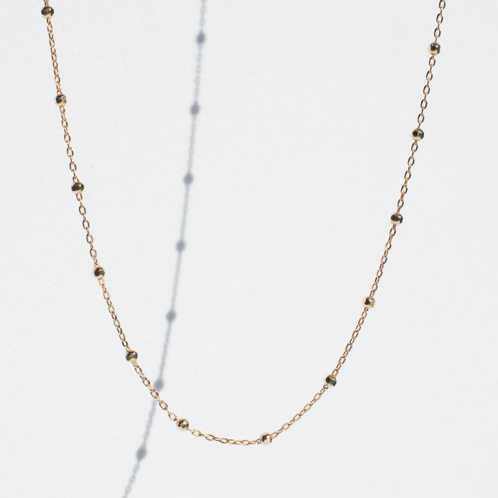 
                      
                        The Beaded Chain by Pear, a delicate gold chain necklace adorned with small faceted beads evenly spaced along its length, is displayed against a light background. Shadows of the necklace and beads, secured by a lobster clasp, are also visible on the backdrop.
                      
                    