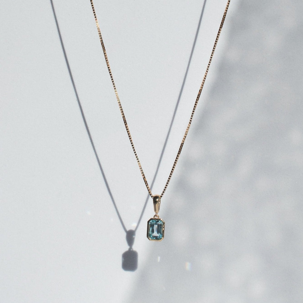 
                      
                        The Estelle Necklace by Pear showcases a delicate gold chain adorned with a rectangular aquamarine gemstone pendant, set impeccably in a solid gold bezel. The necklace and pendant cast soft shadows on a white background, enhancing the elegant composition.
                      
                    