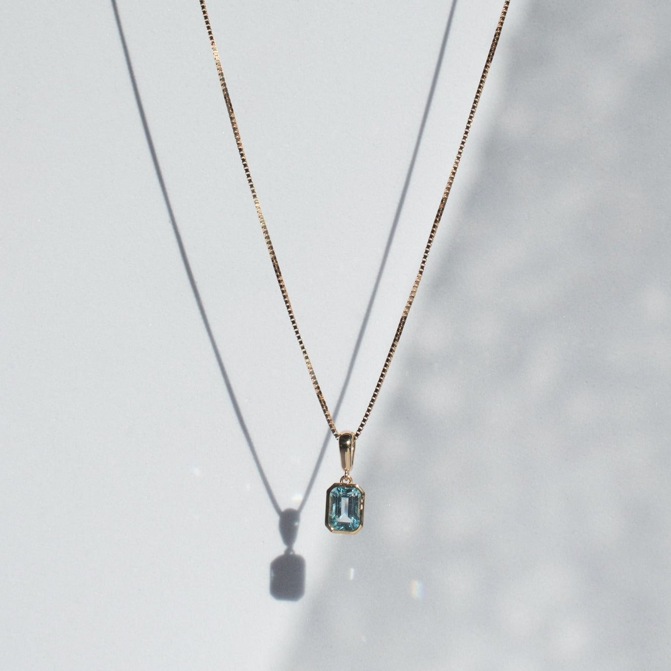 The Estelle Necklace by Pear showcases a delicate gold chain adorned with a rectangular aquamarine gemstone pendant, set impeccably in a solid gold bezel. The necklace and pendant cast soft shadows on a white background, enhancing the elegant composition.