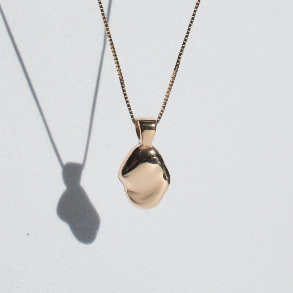 
                      
                        A small, smooth, hand-carved oval pendant of the Ophelia Necklace by Pear hangs from a thin, elegant solid gold chain against a plain light background. The pendant casts a shadow, adding depth to the composition.
                      
                    