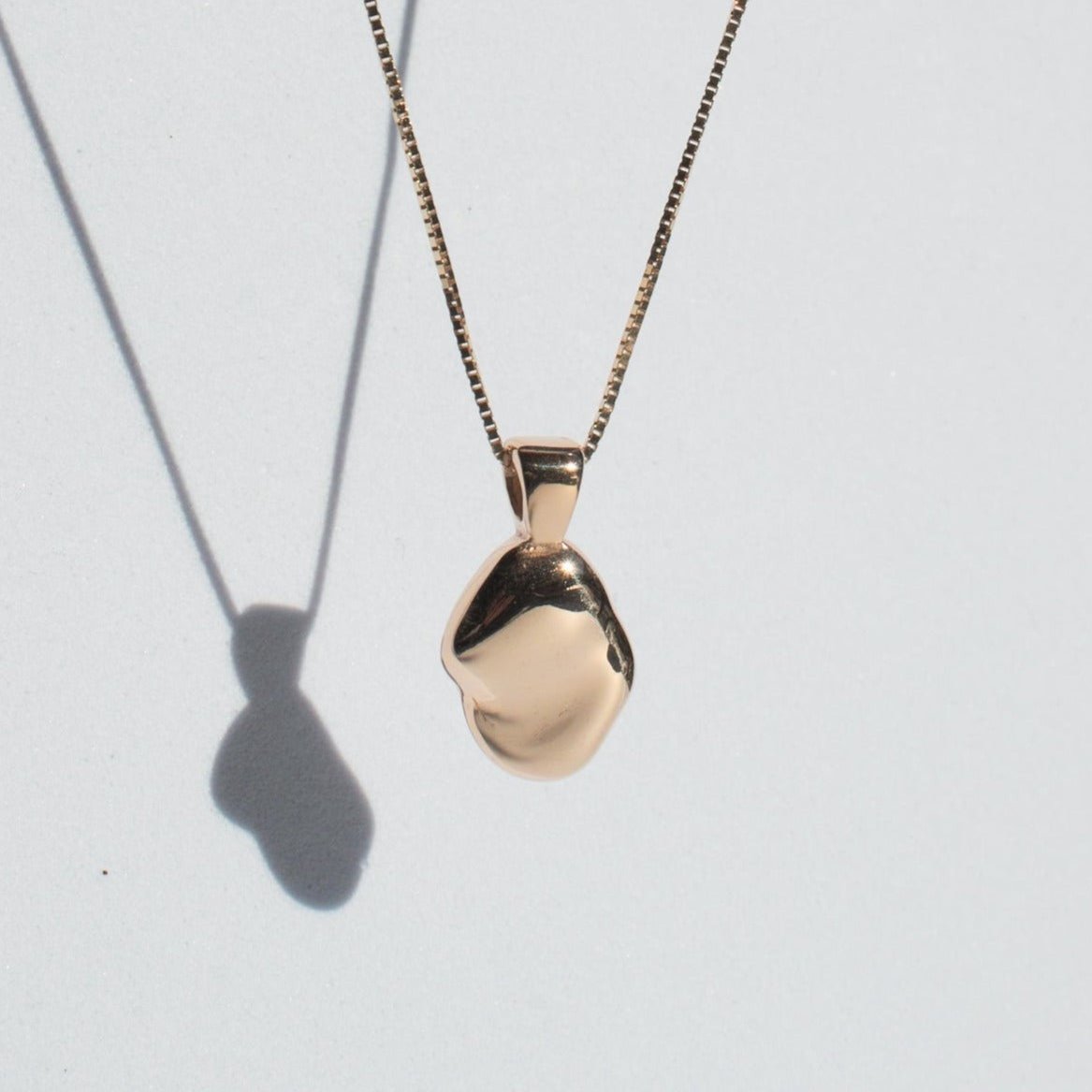 A small, smooth, hand-carved oval pendant of the Ophelia Necklace by Pear hangs from a thin, elegant solid gold chain against a plain light background. The pendant casts a shadow, adding depth to the composition.