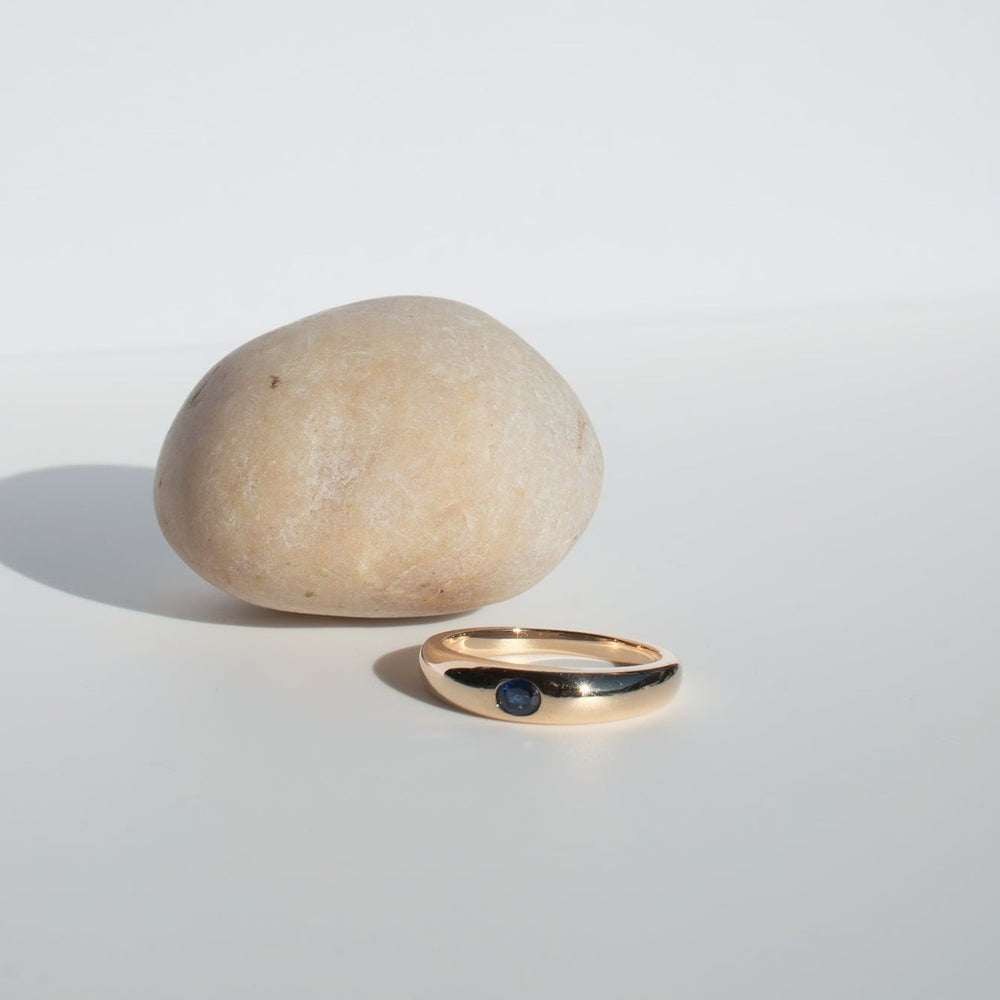 
                      
                        Placed on a white surface against a light background, the Sapphire Dome Ring by Pear, featuring a dark blue sapphire gemstone set in 14K gold, sits elegantly before a smooth beige stone. The ring casts a soft shadow as it rests in front of the stone.
                      
                    