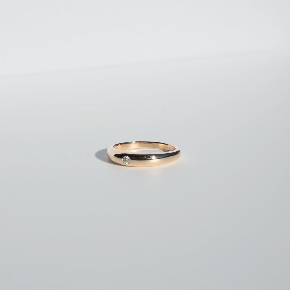 
                      
                        A minimalist and elegant Diamond Dome Ring by Pear, featuring a simple gold band adorned with a small 2mm diamond, rests on a plain, light grey surface.
                      
                    