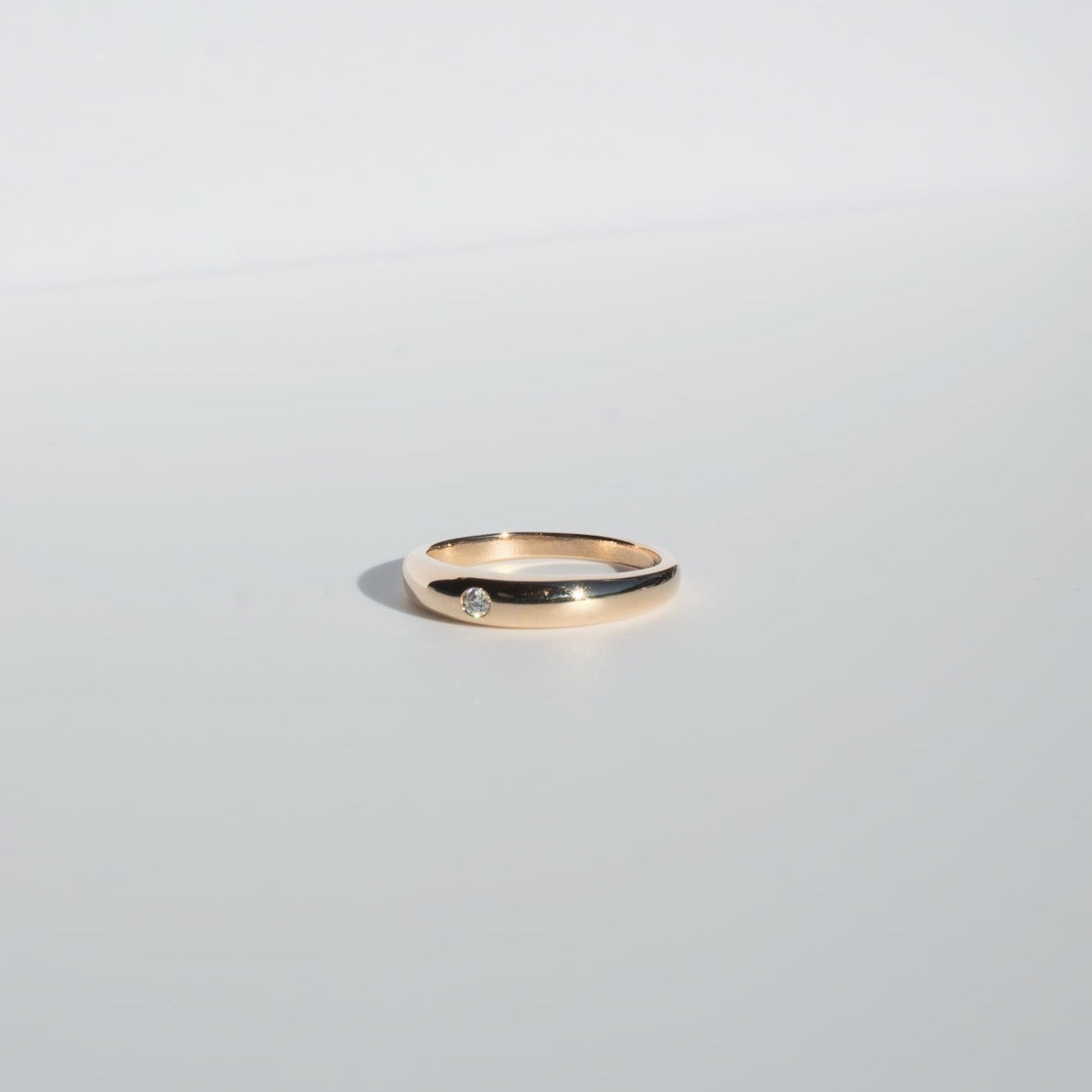 A minimalist and elegant Diamond Dome Ring by Pear, featuring a simple gold band adorned with a small 2mm diamond, rests on a plain, light grey surface.