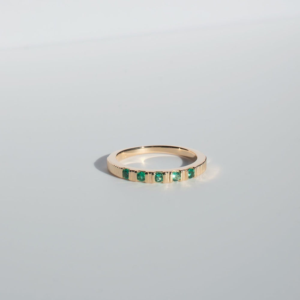 
                      
                        The Pear Gemstone Ribbed Ring, a solid gold band adorned with a row of small, evenly spaced green gemstones, is showcased against a light, neutral background. The ring exudes a subtle shine that highlights its polished surface and the vibrant hue of the stones along with its intricate ribbed design.
                      
                    