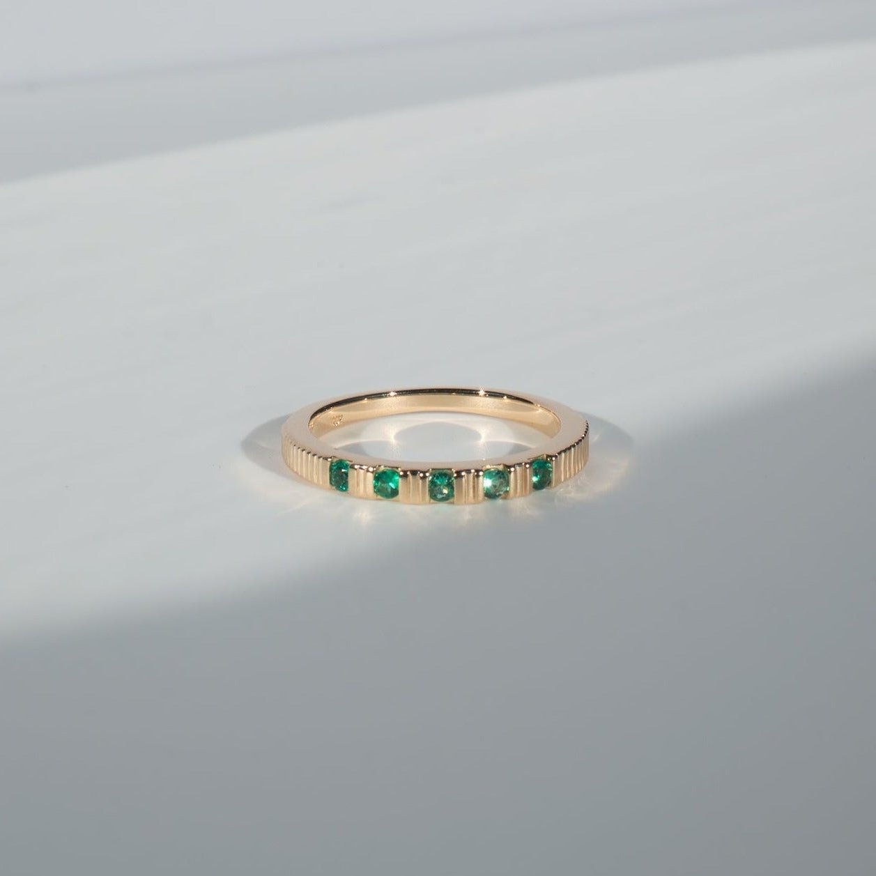 A delicate gold ring set with small, evenly spaced emerald stones is displayed on a light gray surface. Shadows softly fall across the surface, emphasizing the ring's subtle shine and the deep green color of the emeralds.