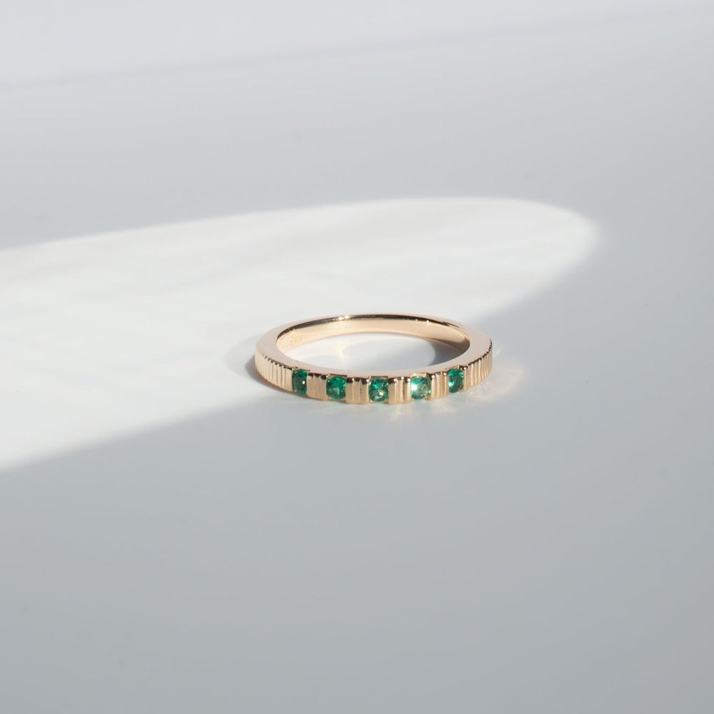 
                      
                        The Pear Gemstone Ribbed Ring, with its minimalist solid gold band adorned with a row of small, evenly spaced green gemstones on its upper side, is showcased on a plain, light-colored surface. Soft natural light illuminates the ring, casting a delicate shadow.
                      
                    