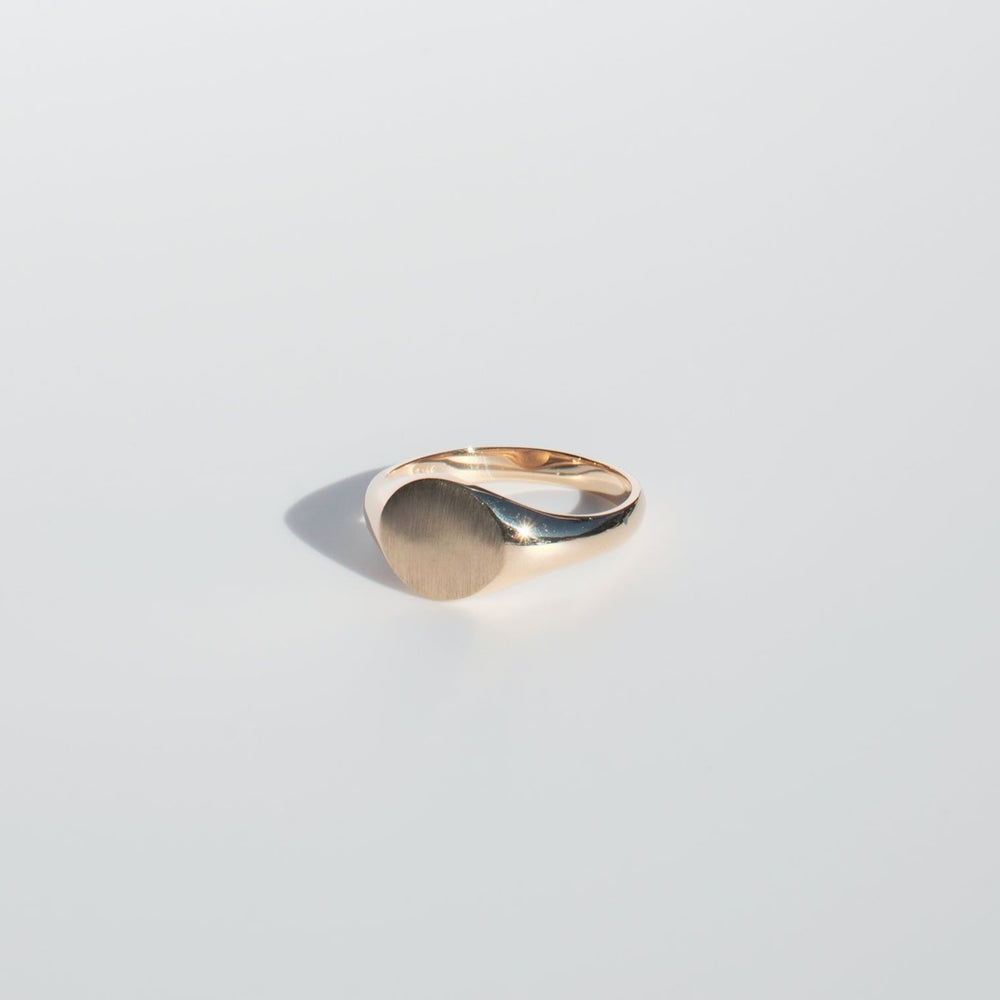 
                      
                        A close-up image showcases the Pear Petite Round Signet Ring, highlighting its minimalist gold design set against a plain, light background. The ring features a smooth, polished band and a flat, circular face ideal for an engraved monogram.
                      
                    