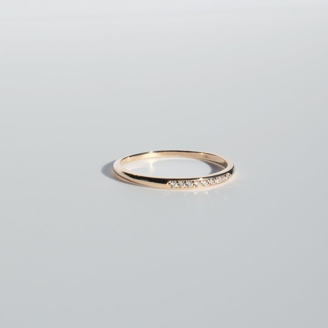 The Esme Diamond Ring by Pear, a thin gold band with a minimalist design, lies on a reflective surface. This exquisite ring showcases a row of small, sparkling diamonds embedded into the band. The background is a smooth, light gray.