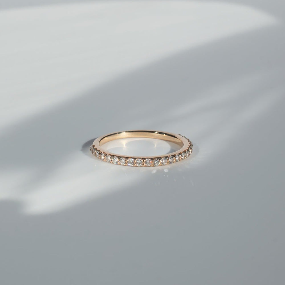 
                      
                        The Pear. Diamond French Band, a delicate gold piece encrusted with small, sparkling diamonds, is resting on a reflective surface and casting a subtle shadow. The background features a soft gradient of light gray and white.
                      
                    