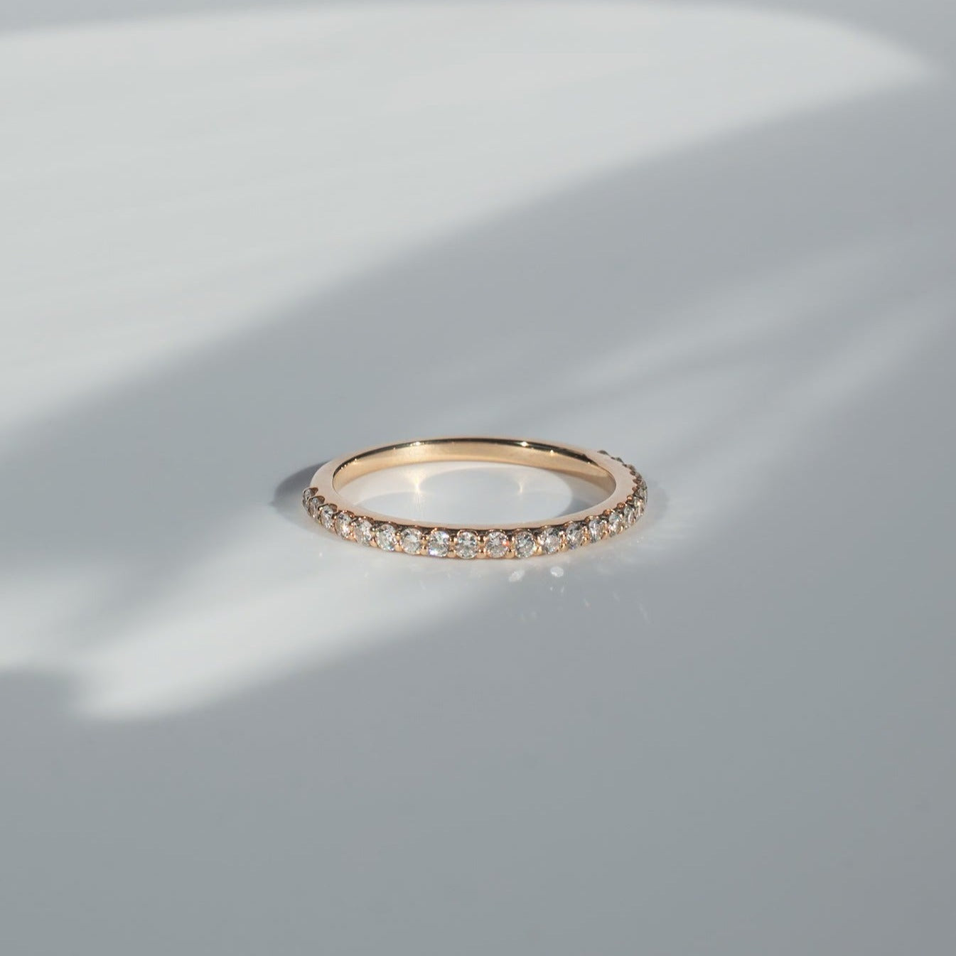 The Pear. Diamond French Band, a delicate gold piece encrusted with small, sparkling diamonds, is resting on a reflective surface and casting a subtle shadow. The background features a soft gradient of light gray and white.