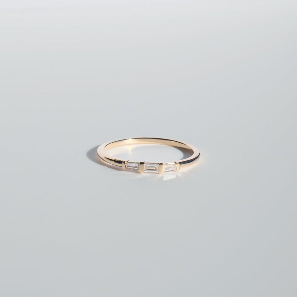 
                      
                        The Edith Diamond Ring by Pear features a minimalist gold design adorned with three small baguette diamonds set in a row along the band. It is showcased against a plain, soft grey background, emphasizing its delicate and elegant aesthetic.
                      
                    