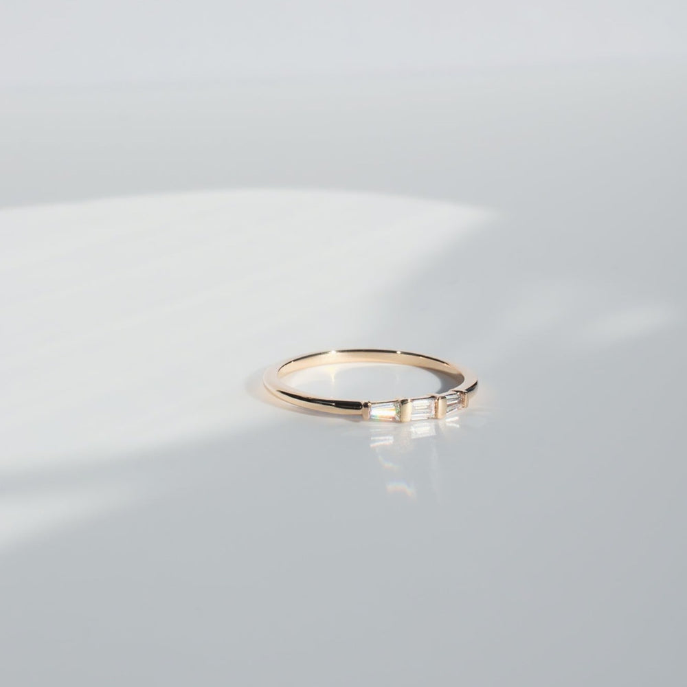 
                      
                        A thin Edith Diamond Ring by Pear, featuring three small baguette diamonds, rests on a smooth, reflective white surface. Soft shadows and light create a serene background, accentuating the ring's elegant and minimalist design.
                      
                    