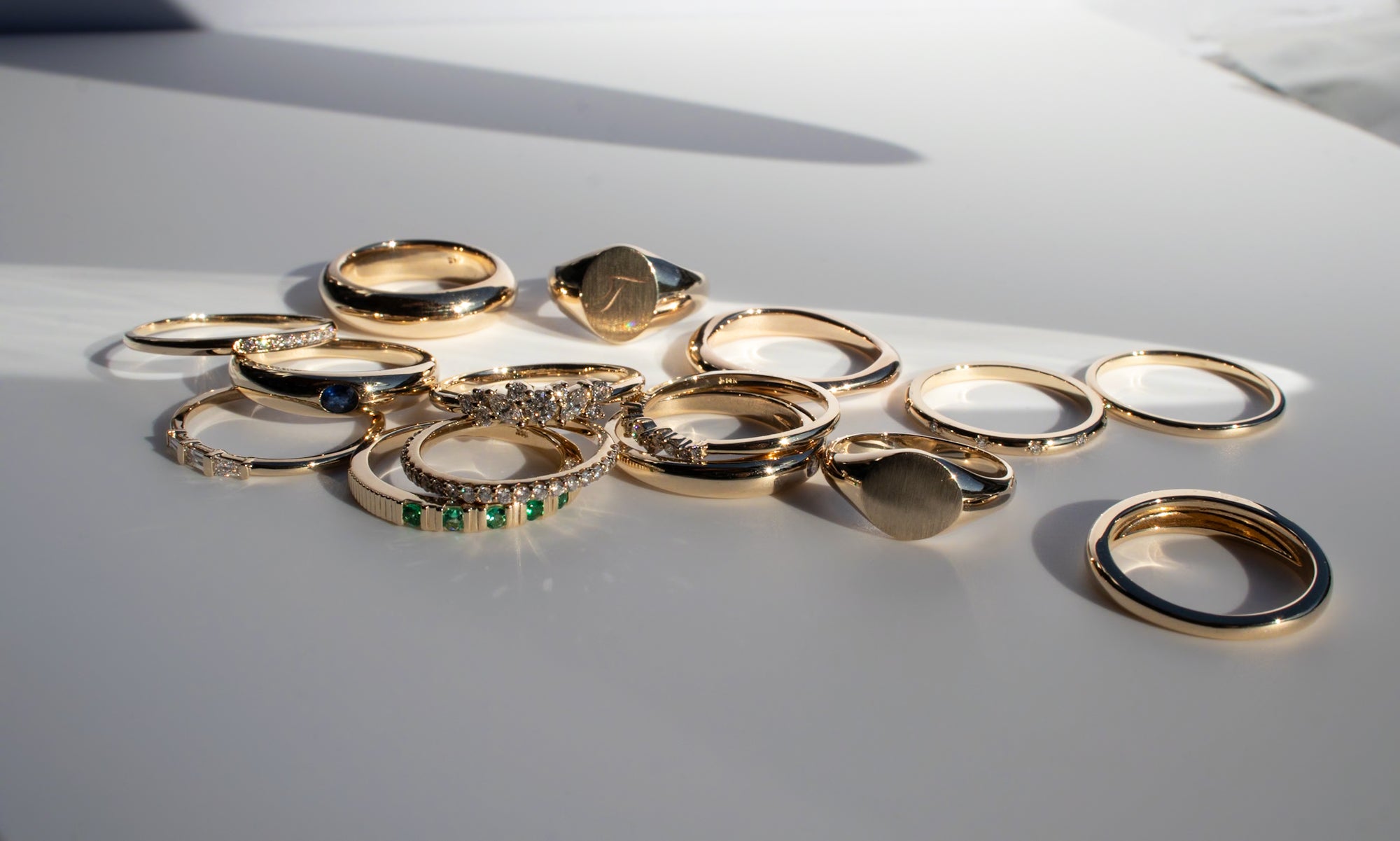 A scattered collection of gold rings lies on a white surface. The rings vary in design, some featuring gemstones in different colors, while others are simple and unadorned bands. The light casts soft shadows, emphasizing the luster of the gold.