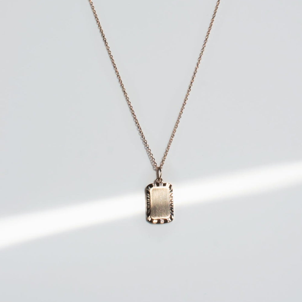 
                      
                        The Engraveable Rectangle Necklace by Pear features a delicate gold pendant with a rectangular charm, elegantly suspended against a plain white background. The fine, length-adjustable cable chain enhances the simplicity and elegance of this piece. A soft beam of light subtly illuminates part of the necklace.
                      
                    