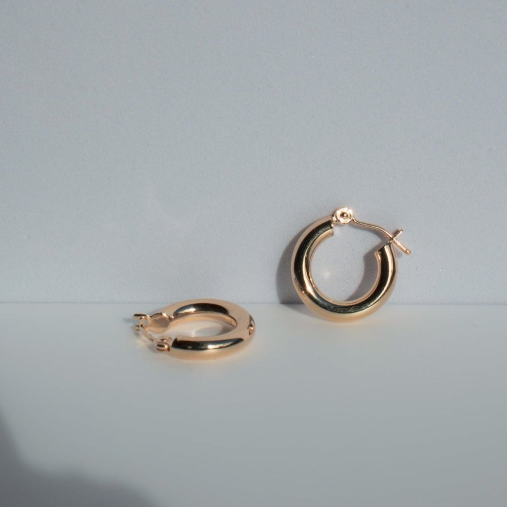 
                      
                        The Pear Mini Thick Hoops, crafted in 14K gold, are displayed against a light gray background. One earring stands upright while the other lies flat. The minimalist scene is well-lit, highlighting the shiny, polished surface of these ideal everyday wear pieces.
                      
                    