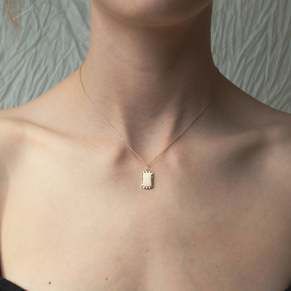 
                      
                        Close-up of a person wearing the Pear Engravable Rectangle Necklace. The delicate gold necklace features a small, rectangular pendant perfect for engraving. The person is dressed in a strapless top, with lightly textured white fabric in the background. The length-adjustable cable chain adds versatility to this elegant piece.
                      
                    