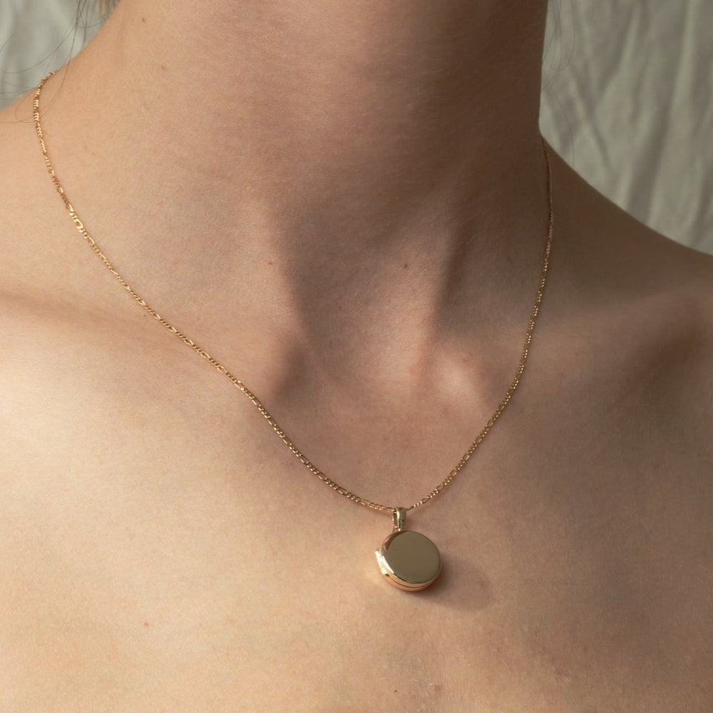 A person wearing the elegant Pear Round Locket, a delicate gold chain necklace featuring a plain 14K gold locket pendant. The neutral background brings focus to the beautiful jewelry, while the visible clavicle and neck enhance the piece's elegance.