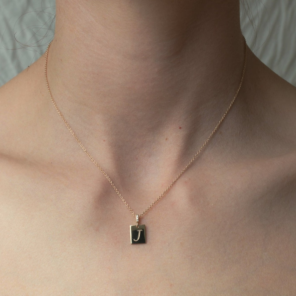 
                      
                        A close-up photograph of a person's neck, wearing the delicate Diamond Cleo Initial Necklace by Pear, featuring a small, square black pendant with a gold letter "J" in its center. The background is neutral and somewhat out of focus. This versatile accessory adds an elegant touch to any outfit.
                      
                    