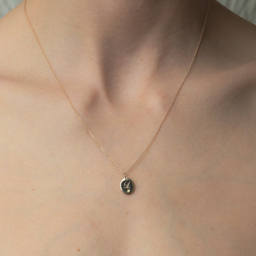 A close-up image captures a person's neck and collarbone adorned with the Pear Oval Engravable Necklace. The delicate gold pendant, featuring a small oval charm, hangs from an adjustable cable chain that beautifully displays its simple bird engraving. The softly blurred background complements the image with a neutral color palette.