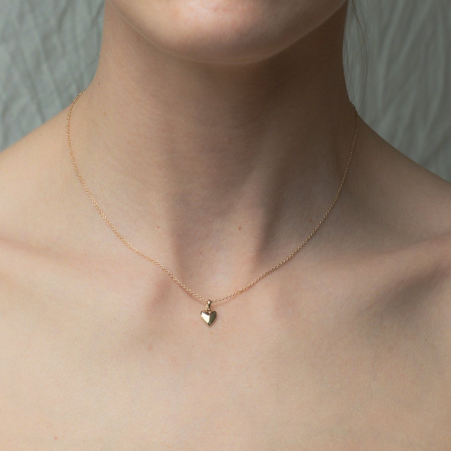 A close-up image of a person wearing the Pear Puffed Heart Necklace showcases its delicate, lightweight design with a small heart-shaped pendant. The background remains neutral, drawing attention to the adjustable cable chain that drapes gracefully around the person's neck and collarbone.