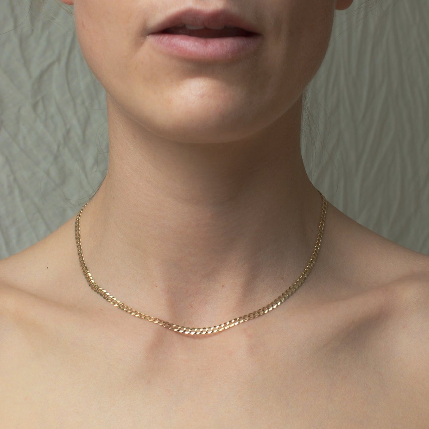 A close-up image shows a person wearing a Pear Curb Chain Necklace made of 14K Solid Yellow Gold around their neck. The background appears to be a textured, light-colored surface. The person's shoulders and part of their mouth are visible, with natural lighting highlighting the necklace's daily wearability.