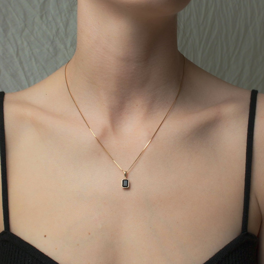 A person wearing the Estelle Necklace by Pear, featuring a small square pendant encased in a solid gold bezel. They are dressed in a black spaghetti strap top, with the background appearing to be a textured, light-colored fabric.