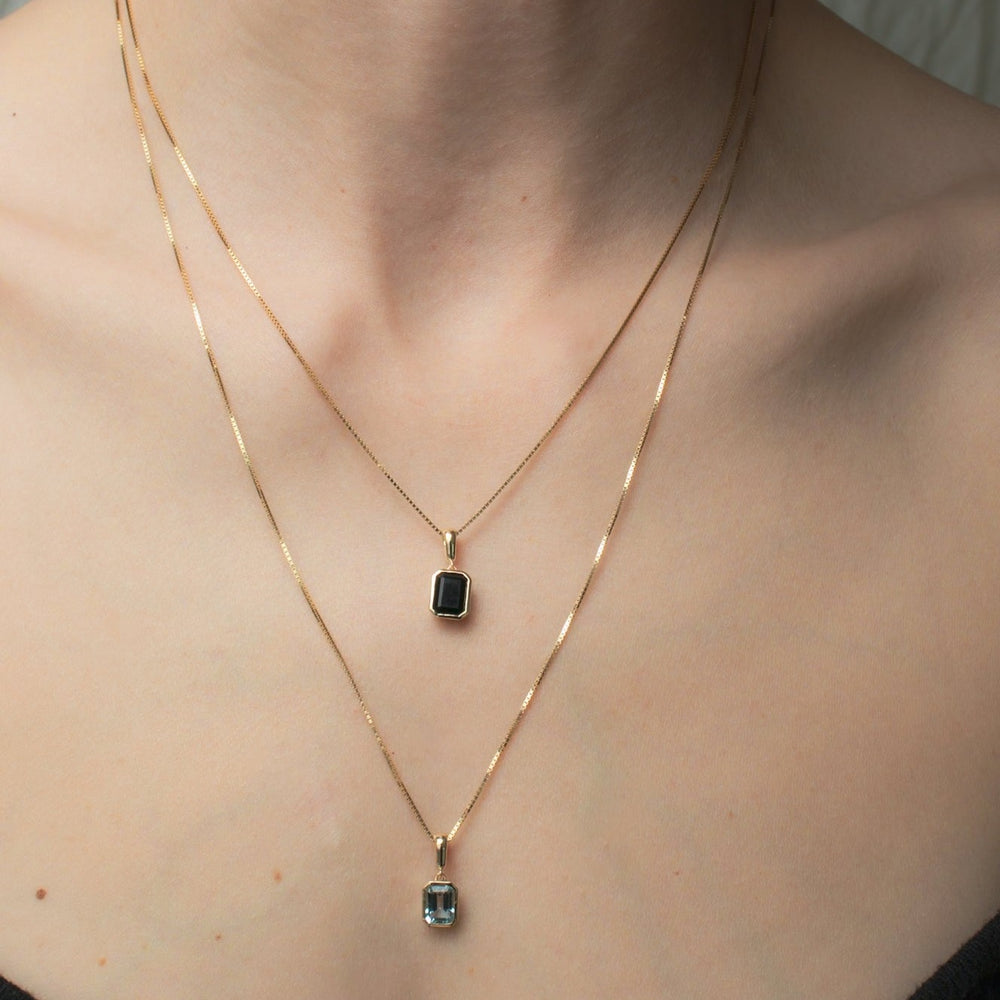 
                      
                        Close-up of a person wearing two Estelle Necklaces by Pear, each adorned with thin gold chains and small rectangular pendants. The upper pendant showcases a black onyx set in a solid gold bezel, while the lower pendant is clear. The focus is on the jewelry against the person's skin, highlighting their neck and collarbone.
                      
                    