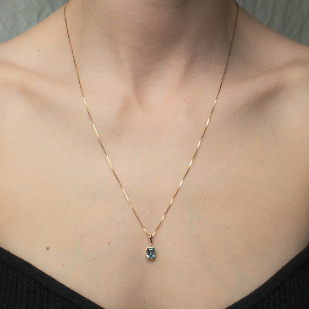 
                      
                        A person wearing the Estelle Necklace by Pear, featuring a square-shaped sky blue topaz pendant set in a solid gold bezel on a thin gold chain. The individual is dressed in an off-the-shoulder black top.
                      
                    