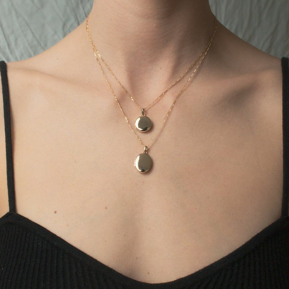 A close-up of a person's collarbone and neck area wearing a black sleeveless top. They are adorned with two delicate gold chain necklaces from Pear, layered elegantly around their neck. One necklace features a Figaro chain with custom engraving, each one boasting a small round pendant from Pear's Round Locket collection that rests at different lengths on the chest.