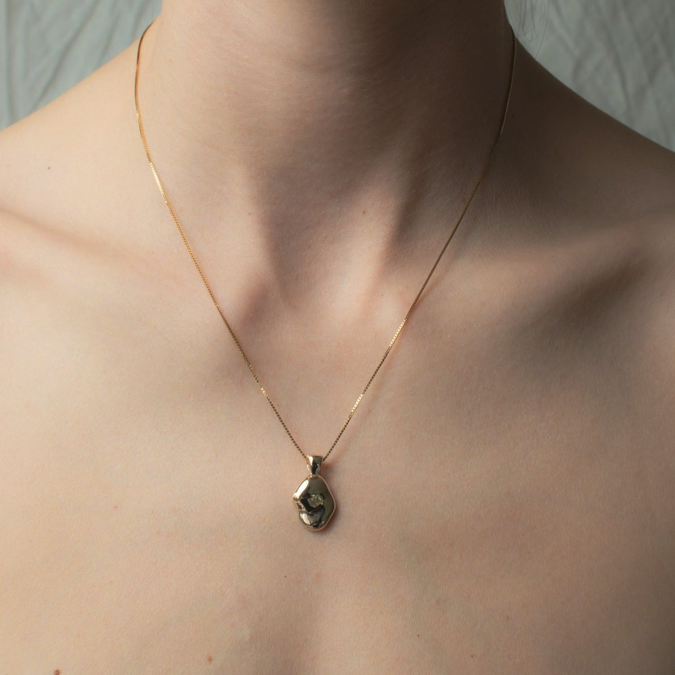 An intimate close-up captures a person's neck graced by the Ophelia Necklace from Pear, a delicate solid gold chain featuring a uniquely, hand-carved oval pendant. The softly blurred background enhances focus on this exquisite piece of jewelry.