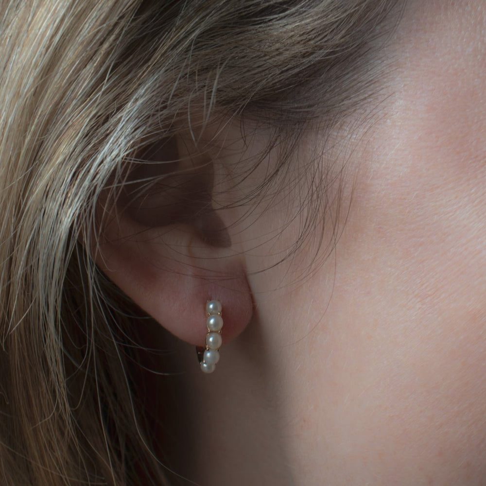 
                      
                        Close-up of a person's ear wearing Pearl Huggies by Pear, featuring a small hoop earring adorned with several freshwater pearls. The skin and ear detail are clearly visible, with light brown hair partially framing the ear. The image showcases the delicate and elegant design of the 14K gold earring.
                      
                    