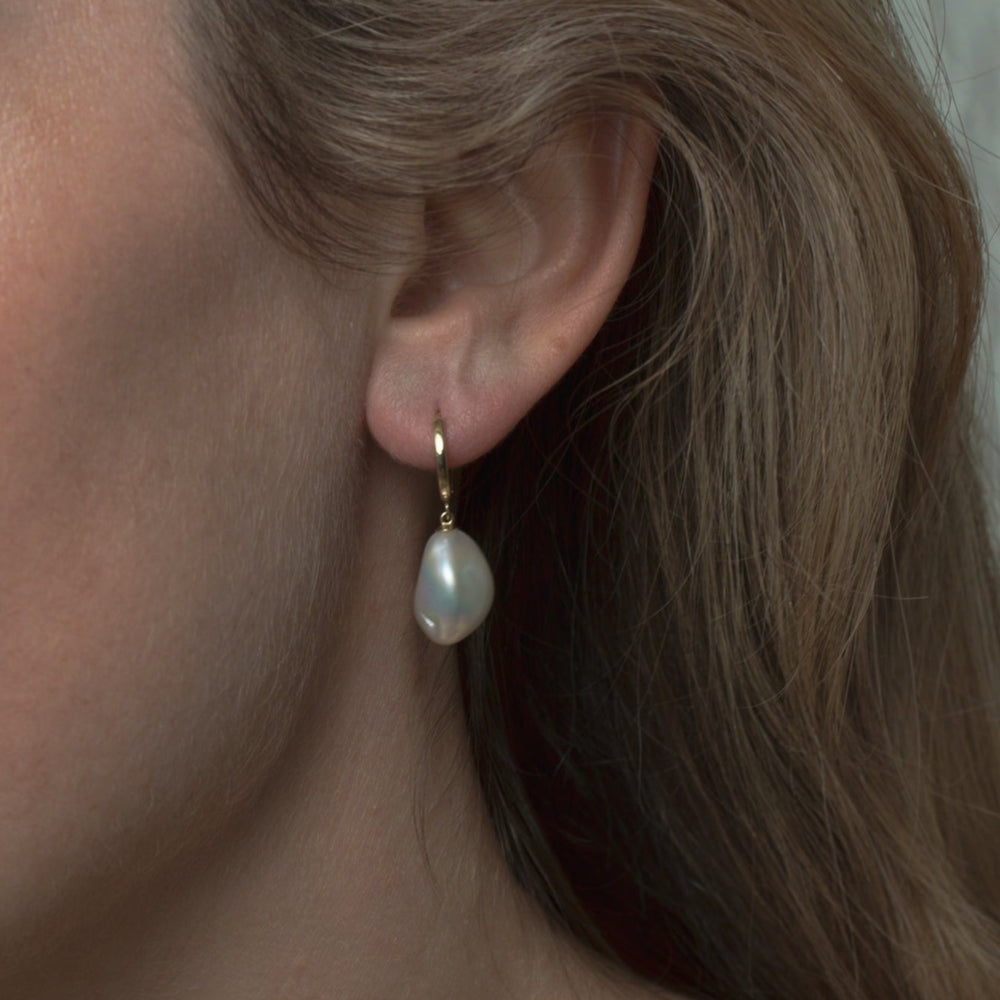 Close-up of a person’s ear wearing a single, elegant Pearl Accented Hoops by Pear, featuring a 14K gold hoop earring with a white teardrop-shaped freshwater Keshi pearl pendant. The person's defined ear and earring are in focus, set against light brown hair and fair skin, epitomizing luxury jewelry.