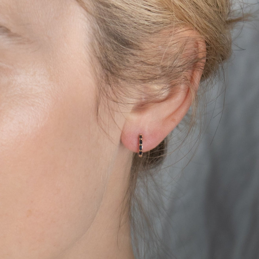 
                      
                        Close-up of a person's ear adorned with Pear's Sapphire Huggies, small gold hoop earrings embedded with baguette sapphires. The ear and earrings stand out against a softly blurred, neutral background. The person's blonde hair is tied back, partially concealing the top of the ear.
                      
                    