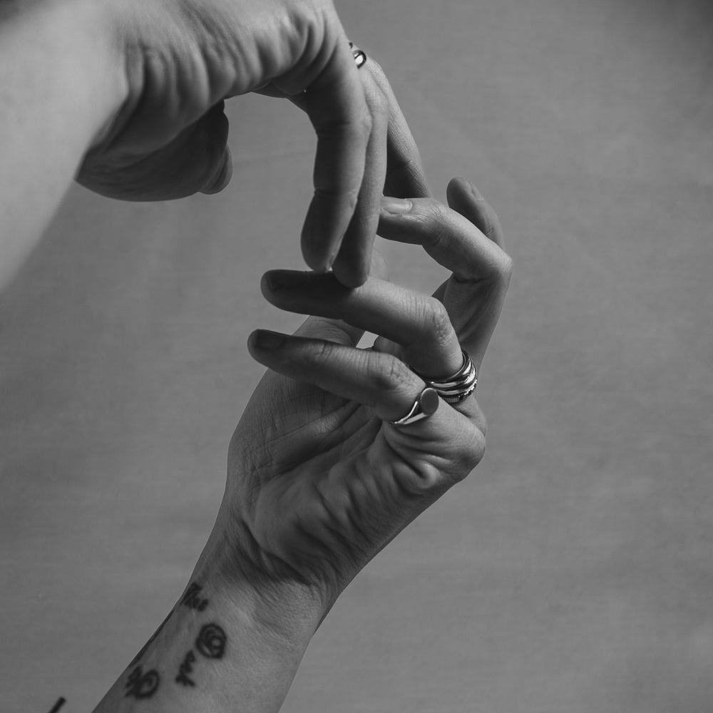 Black and white photo of two hands, each adorned with rings and bracelets. One wrist has a tattoo of small symbols. The hands are posed delicately against a plain background.