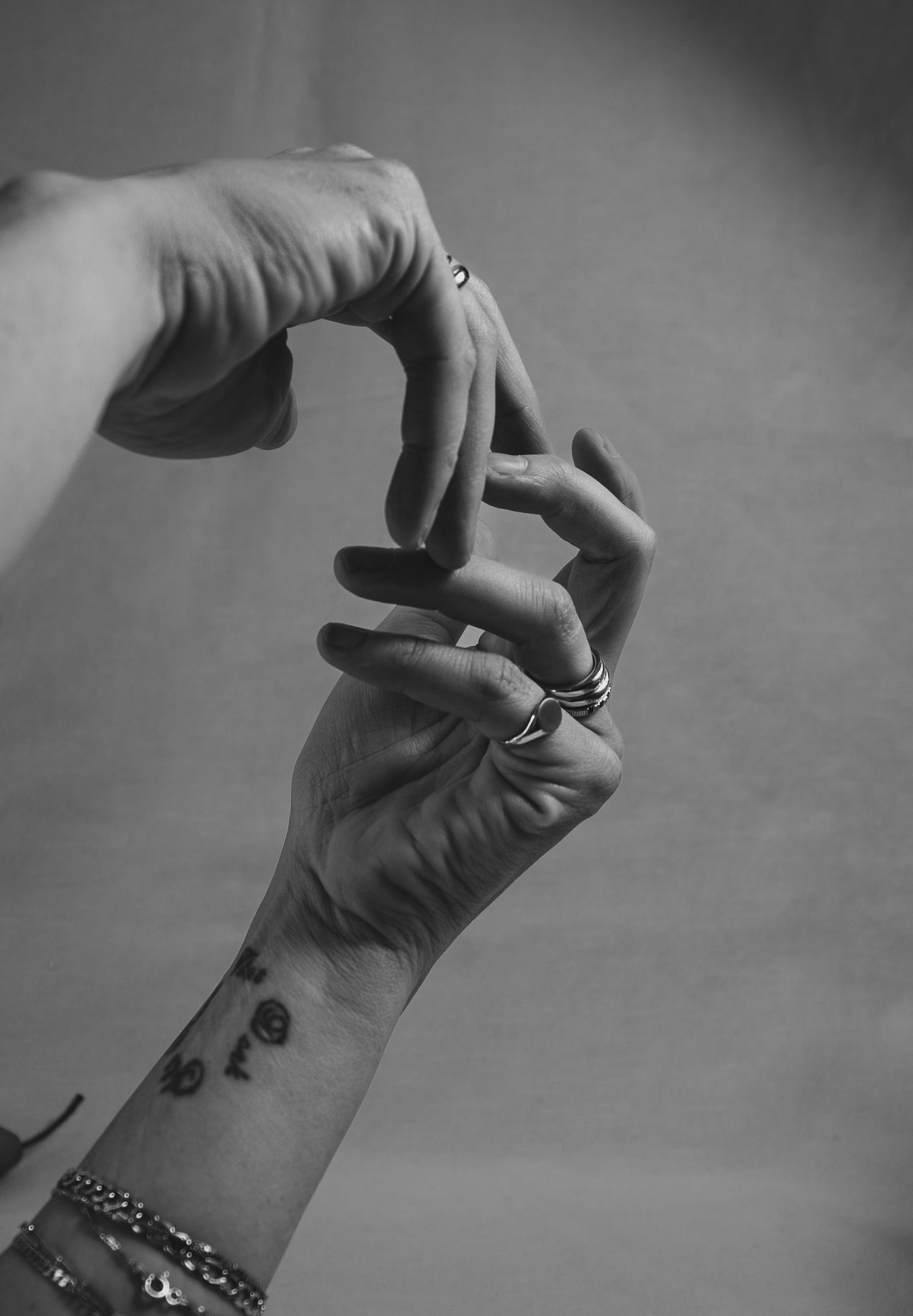 Black and white photo of two hands, each adorned with rings and bracelets. One wrist has a tattoo of small symbols. The hands are posed delicately against a plain background.